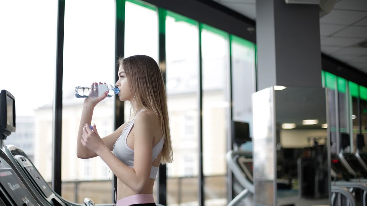 A young woman drinks water during her workout on a treadmill at a modern indoor gym.